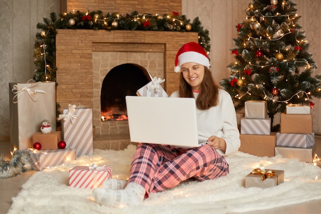 Foto de uma linda garota encantadora atraente trabalhando na véspera do ano novo com seu laptop ou tendo uma vídeo chamada para cumprimentar alguém, vestindo calças xadrez e chapéu de Papai Noel, olha para a tela do caderno com um sorriso.