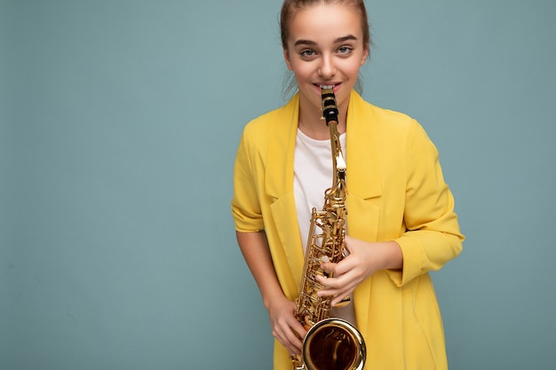 Foto de uma linda garotinha morena feliz e sorridente, vestindo uma elegante jaqueta amarela, isolada sobre uma parede de fundo azul tocando saxofone, olhando para a câmera