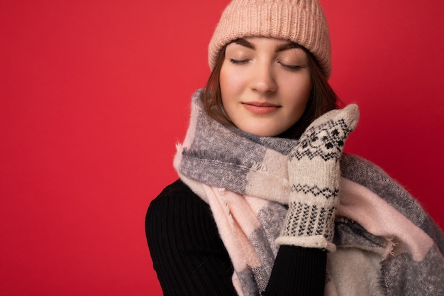 Foto de uma linda jovem morena feliz fofa isolada sobre uma parede de fundo vermelho, usando luvas de lenço de inverno e um chapéu quente, apreciando com os olhos fechados