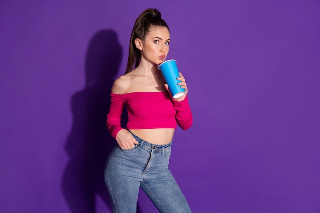 Foto de uma linda jovem sonhadora segurar copo braço bolso lábios canudo bebida refrigerante usar jeans rosa top isolado fundo de cor violeta