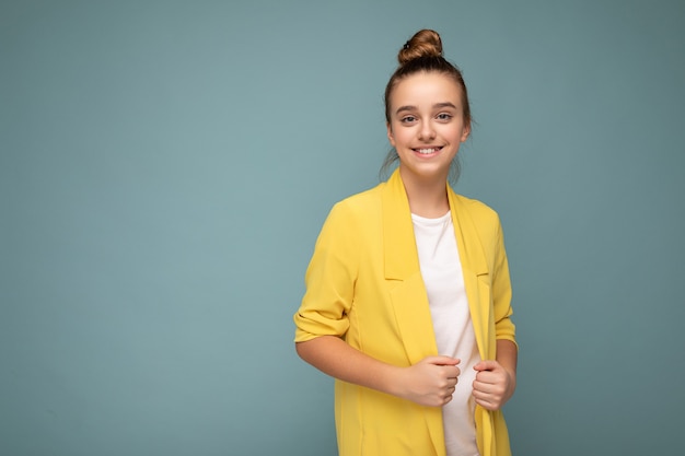 Foto de uma linda menina morena feliz e sorridente, vestindo uma jaqueta amarela da moda e uma camiseta branca isolada sobre a parede de fundo azul, olhando para a câmera