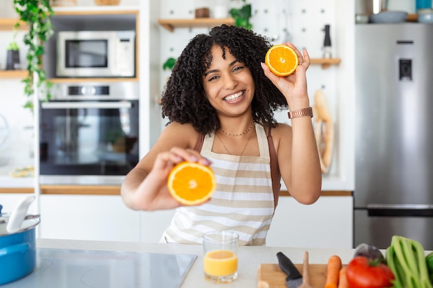 Foto de uma linda mulher afro-americana sorrindo e segurando duas partes de laranja enquanto cozinha salada de legumes no interior da cozinha em casa