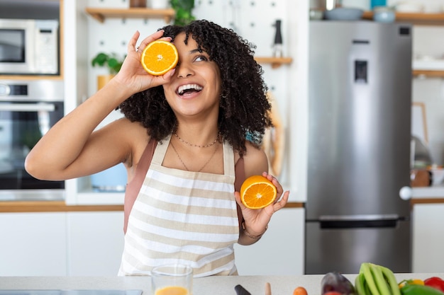 Foto de uma linda mulher afro-americana sorrindo e segurando duas partes de laranja enquanto cozinha salada de legumes no interior da cozinha em casa