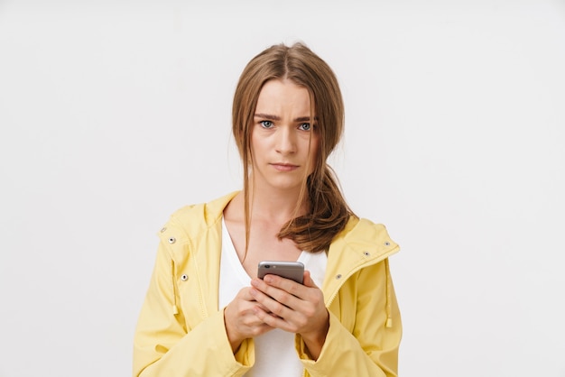 Foto de uma linda mulher confusa em uma capa de chuva usando o celular e olhando para a câmera