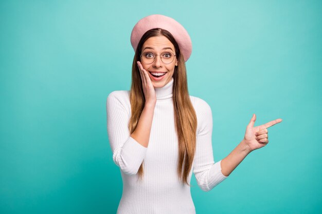 Foto de uma linda senhora chocada com o dedo direto do lado do espaço vazio apresentando banner de anúncio de venda mão na bochecha especificações de desgaste boina rosa boné de gola alta branco isolado fundo de cor verde-azulado