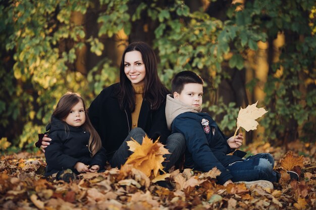 Foto de uma mãe com longos cabelos negros em um casaco preto, um lindo garotinho com sua irmã mais nova segurando buquês de folhas de outono