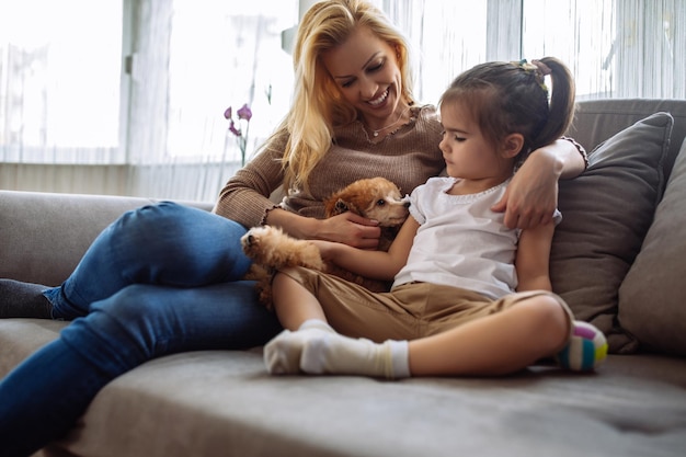 Foto de uma mãe e filha sentadas em um sofá com um cachorrinho fofo