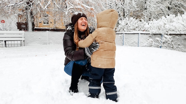 Foto de uma mãe sorridente com um casaco com seu filho fofo em uma jaqueta bege se divertindo jogando bolas de neve no Playground no Parque