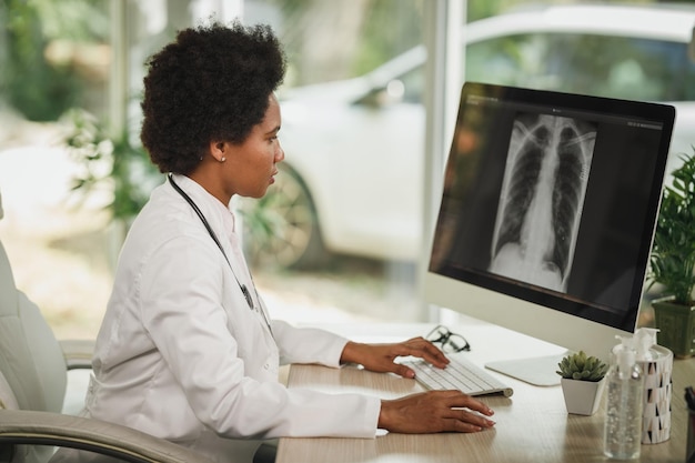 Foto de uma médica africana sentada sozinha em seu consultório e olhando o raio-X no computador durante a pandemia de COVID-19.