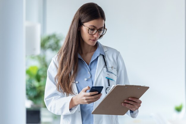 Foto de uma médica bonita usando seu telefone celular enquanto analisa relatórios médicos em uma consulta médica.