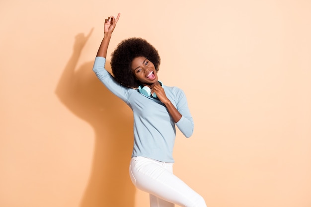 Foto de uma mulher afro-americana apontando um dedo para cima segurando fones de ouvido sem fio isolados em um fundo de cor bege pastel