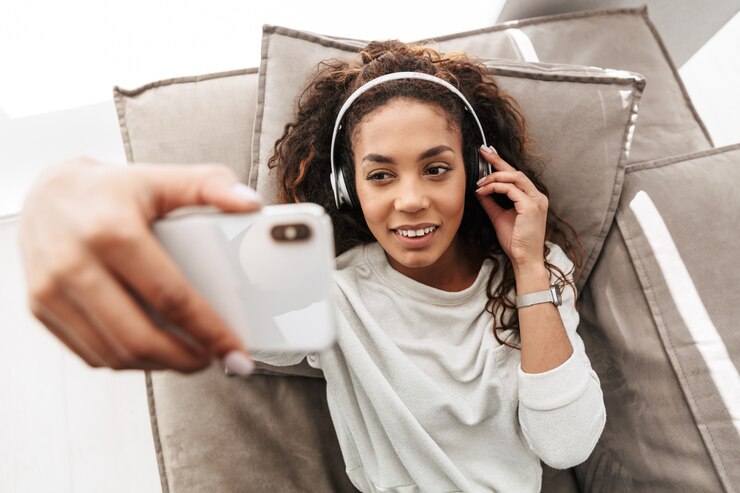 Foto foto de uma mulher afro-americana satisfeita usando fones de ouvido, tirando uma selfie no celular, enquanto estava deitada no sofá em um apartamento bem iluminado