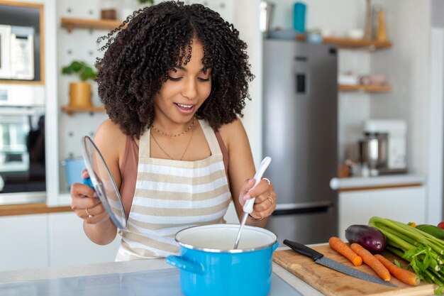 Foto de uma mulher afro-americana segurando colher de concha enquanto cozinha sopa com legumes frescos na cozinha em casa