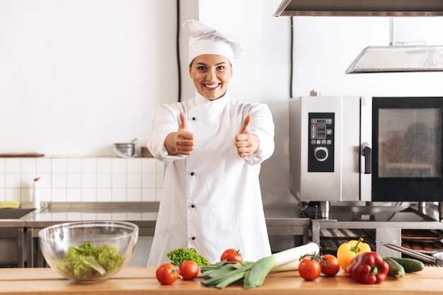 Foto de uma mulher alegre, chef vestindo uniforme branco, cozinhando uma refeição com legumes frescos, na cozinha de um restaurante