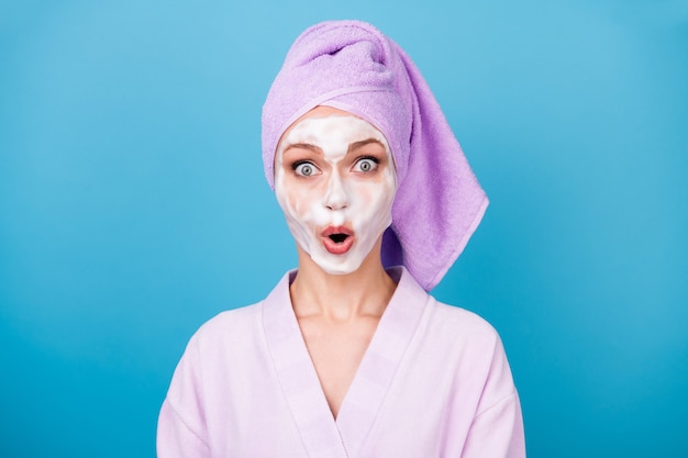 Foto de uma mulher bonita, máscara branca de espuma, boca aberta, usar toalha roxa, turbante, roupão de banho isolado fundo de cor azul