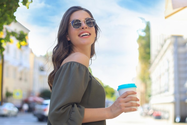 Foto de uma mulher bonita sorridente de vestido verde segurando um copo de papel andando ao ar livre pela rua