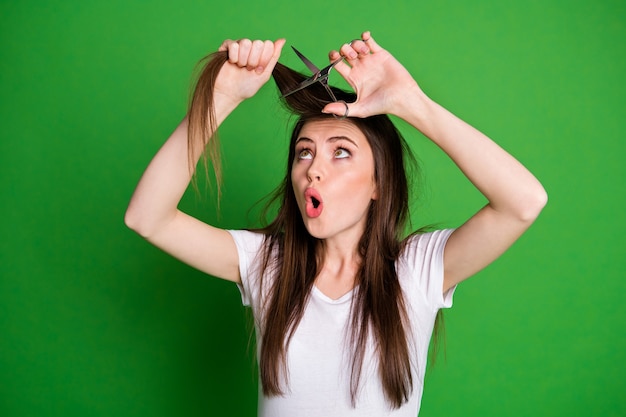 Foto de uma mulher chocada cortando o cabelo com uma tesoura isolada em um fundo de cor verde vívido