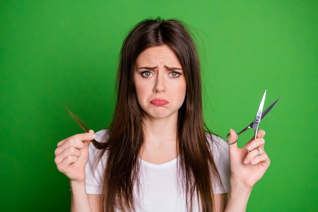 Foto de uma mulher de beicinho triste segurando uma mecha de cabelo em uma tesoura isolada em um fundo de cor verde vívido