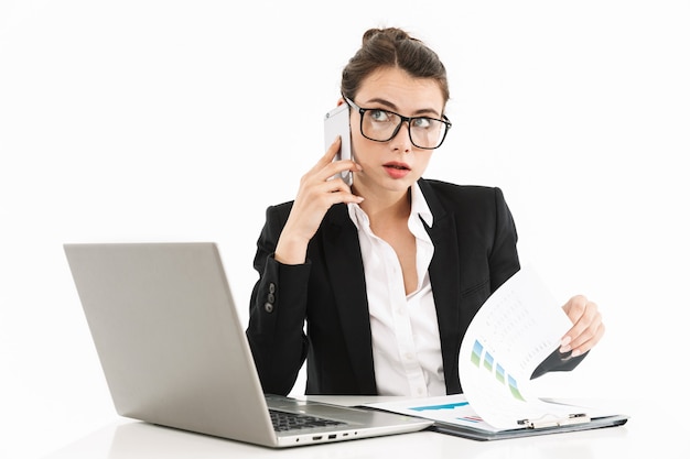 Foto de uma mulher de negócios atraente, vestida com roupa formal, sentada na mesa e trabalhando em um laptop no escritório, isolada sobre uma parede branca