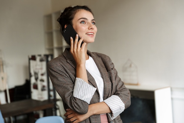 Foto foto de uma mulher de negócios jovem otimista positiva dentro de casa em casa falando por telefone celular.