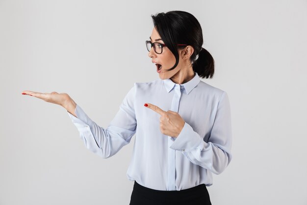 Foto de uma mulher de negócios madura usando óculos em pé no escritório e mostrando copyspace, isolada sobre uma parede branca