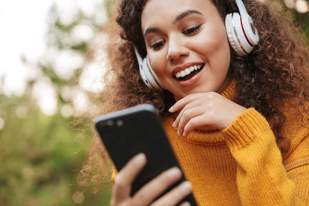 Foto foto de uma mulher encaracolada jovem bonita feliz satisfeita sentar no banco no parque ao ar livre, ouvindo música com fones de ouvido, usando o telefone celular.