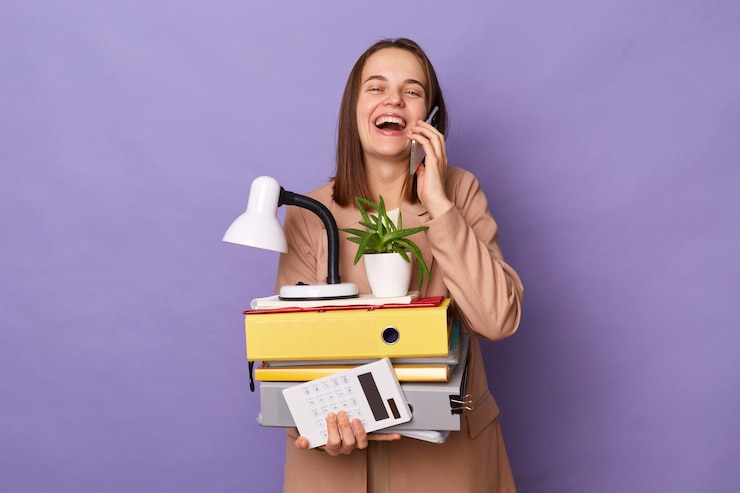 Foto foto de uma mulher feliz rindo vestindo jaqueta bege segurando muitas pastas de documentos isoladas sobre fundo lilás falando no celular ostentando seu novo emprego regozijando-se