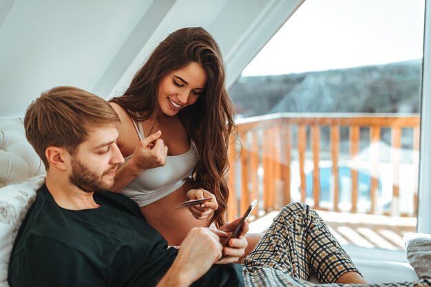 Foto de uma mulher grávida e seu marido deitado na cama e usando o celular