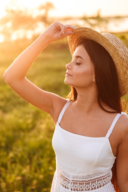 Foto de uma mulher jovem e bonita com longos cabelos escuros em um vestido branco e chapéu de palha, enquanto caminhava na natureza durante um dia ensolarado