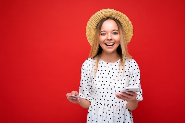 Foto de uma mulher loira atraente e positiva com roupas de verão, usando o celular e segurando um cartão de crédito