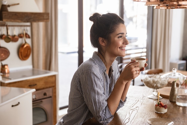 Foto de uma mulher morena feliz tomando café enquanto toma o café da manhã na elegante cozinha de casa