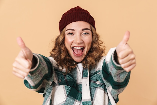 Foto de uma mulher muito alegre com um chapéu de malha rindo e gesticulando com os polegares isolada em bege