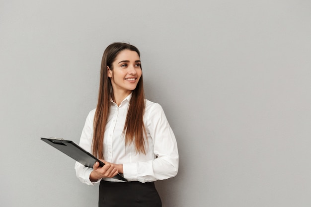 Foto foto de uma mulher profissional de camisa branca e saia preta segurando uma prancheta com documentos no escritório e olhando de lado na copyspace, isolada sobre uma parede cinza