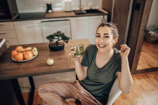 Foto de uma mulher saboreando seu smoothie verde na cozinha em casa. Olhando para a câmera.