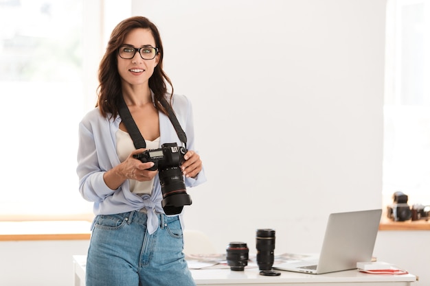 Foto de uma mulher sorridente jovem fotógrafo satisfeito no escritório, segurando a câmera.