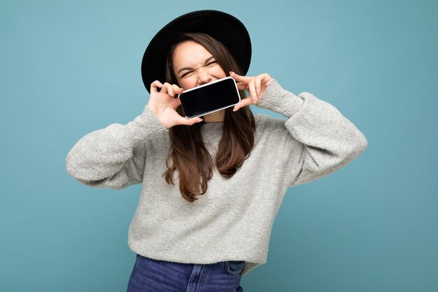 Foto de uma pessoa linda mulher positiva usando chapéu preto e suéter cinza segurando o celular mostrando smartphone isolado no fundo com olhos próximos.