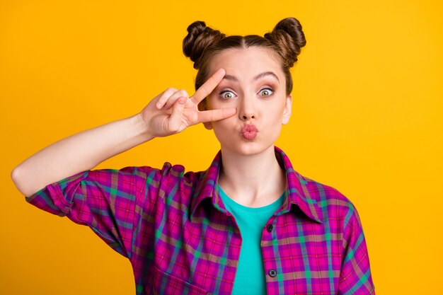 Foto foto de uma senhora adolescente atraente e funky com dois pãezinhos engraçados mostrando o símbolo do sinal de v perto do olho enviando beijos de ar coquete flertando vestir uma camisa xadrez casual isolado fundo de cor amarela brilhante