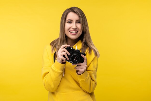 Foto de uma senhora atraente segura a câmera com um sorriso e quer tirar uma foto. Veste casuais com capuz amarelo, fundo de cor amarela isolado.