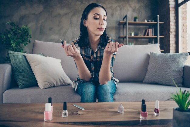 Foto de uma senhora bonita corrigindo as unhas aplicando o esmalte final soprando, ajudando a secar roupas casuais sentadas em um apartamento aconchegante com sofá