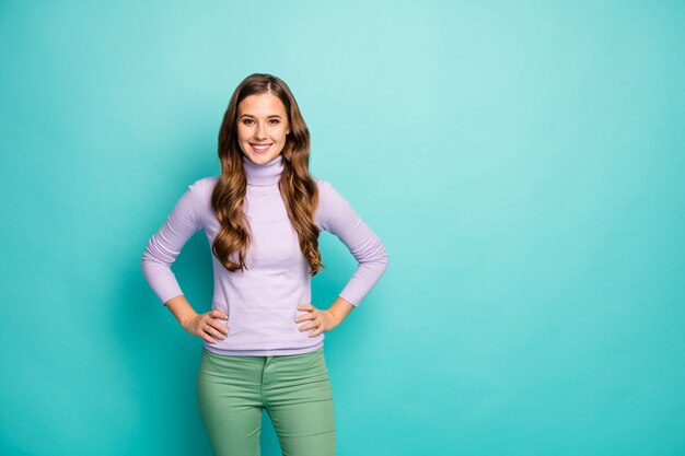 Foto de uma senhora bonita e bonita penteado encaracolado longo, bom humor, mãos aos lados usam suéter roxo lilás calça verde pastel isolada cor azul