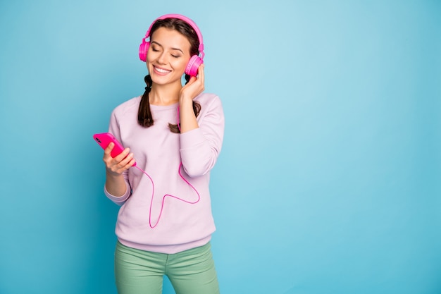 Foto de uma senhora bonita segurando um telefone com fones de ouvido brilhantes nas orelhas ouvindo rádio juvenil, olhos fechados, usar calças casuais elegantes de pulôver rosa isolado cor azul