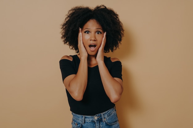 Foto de uma senhora de cabelo encaracolado de pele muito escura chocada, de mãos dadas nas maçãs do rosto, não acredita por causa dos preços baixos de desconto, use uma camiseta preta da moda isolada sobre o fundo bege escuro do estúdio