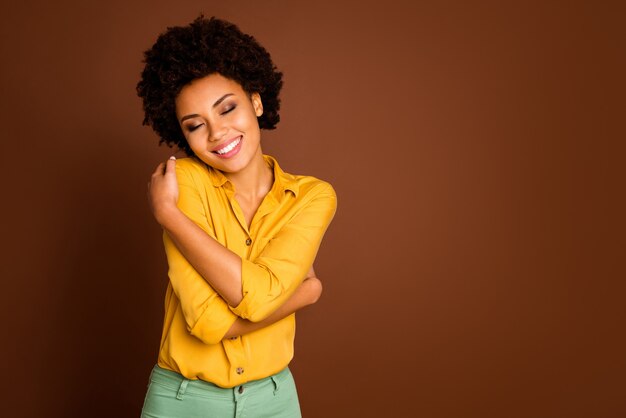 Foto de uma senhora encantadora de pele escura segurando os ombros com os olhos fechados, divirta-se, ame-se, blusa bonita, roupas, camisa amarela, calça verde