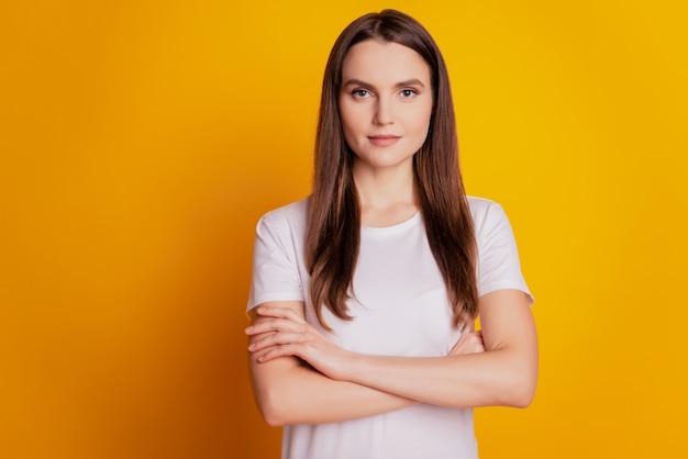 Foto de uma senhora encantadora e confiante com as mãos cruzadas e vestindo uma camiseta branca posando em um fundo amarelo