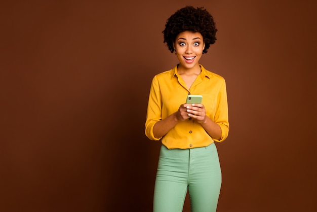 Foto de uma senhora engraçada de pele escura ondulada segurando um telefone influenciador com a boca aberta ler comentários positivos usar camisa amarela calça verde isolada cor marrom