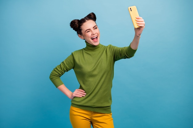 Foto de uma senhora glamour engraçada segurando a mão do telefone fazendo selfies tirando fotos para o blog Os seguidores de olhos piscando usam calças verdes de gola alta amarela com parede de cor azul isolada