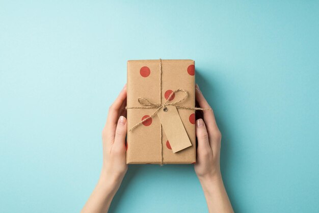 Foto de visão superior em primeira pessoa das decorações do dia dos namorados mãos de uma jovem segurando uma caixa de presente de papel artesanal com padrão de bolinhas e laço de barbante em fundo azul pastel isolado com copyspace