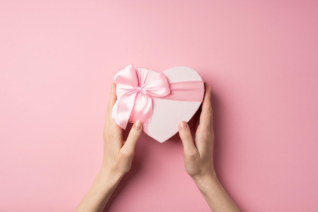 Foto de visão superior em primeira pessoa de decorações de dia dos namorados mãos de jovem segurando caixa de presente em forma de coração com laço de fita rosa em fundo rosa pastel isolado