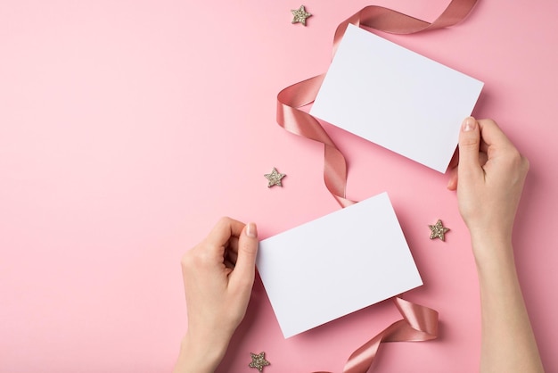 Foto de visão superior em primeira pessoa de decorações de dia dos namorados mãos de mulher segurando duas folhas de papel estrelas brilhantes e fita de cetim cacheada em fundo rosa pastel isolado com espaço em branco