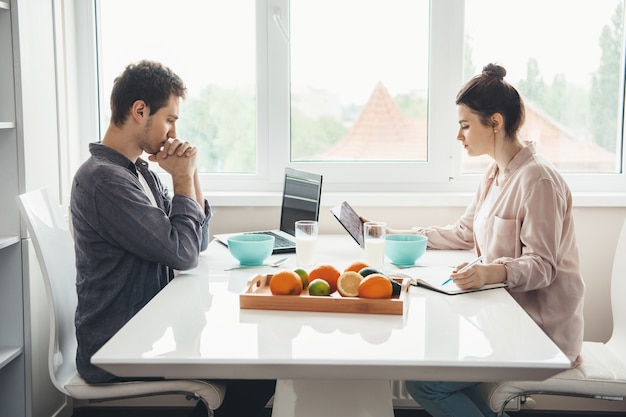 Foto de vista lateral de um casal caucasiano sentado à mesa comendo cereais com leite enquanto trabalhava no laptop